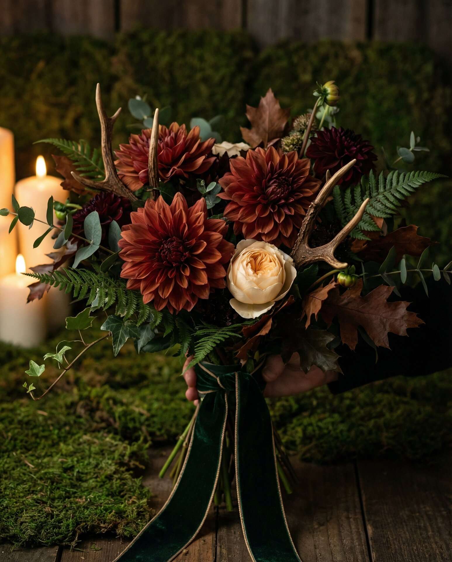 Bridal bouquet of dark dahlias, trailing ivy, deep ferns, antler accents, and a single cream bloom against a deep forest green velvet backdrop
