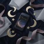 Midnight Celestial wedding invitation on a phone screen, surrounded by gold crescent moons and stars on grey silk