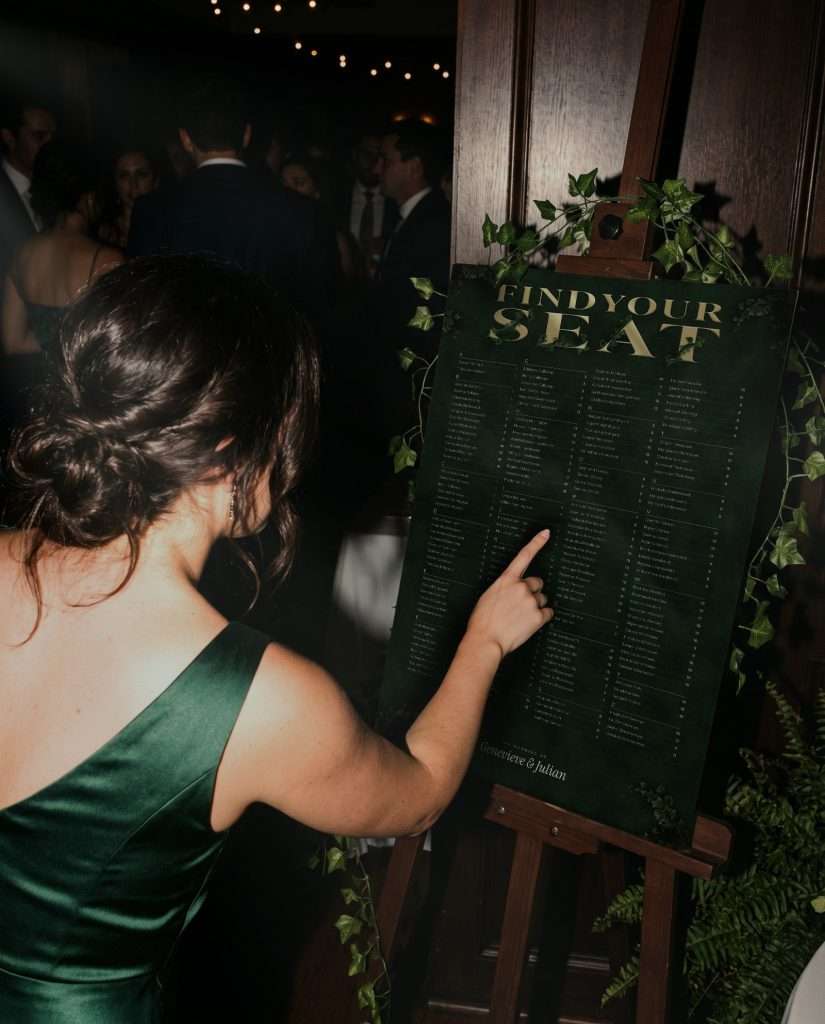 A guest pointing to her name on the Enchanted Forest seating chart on an easel at a candlelit reception, from the Day-Of Suite