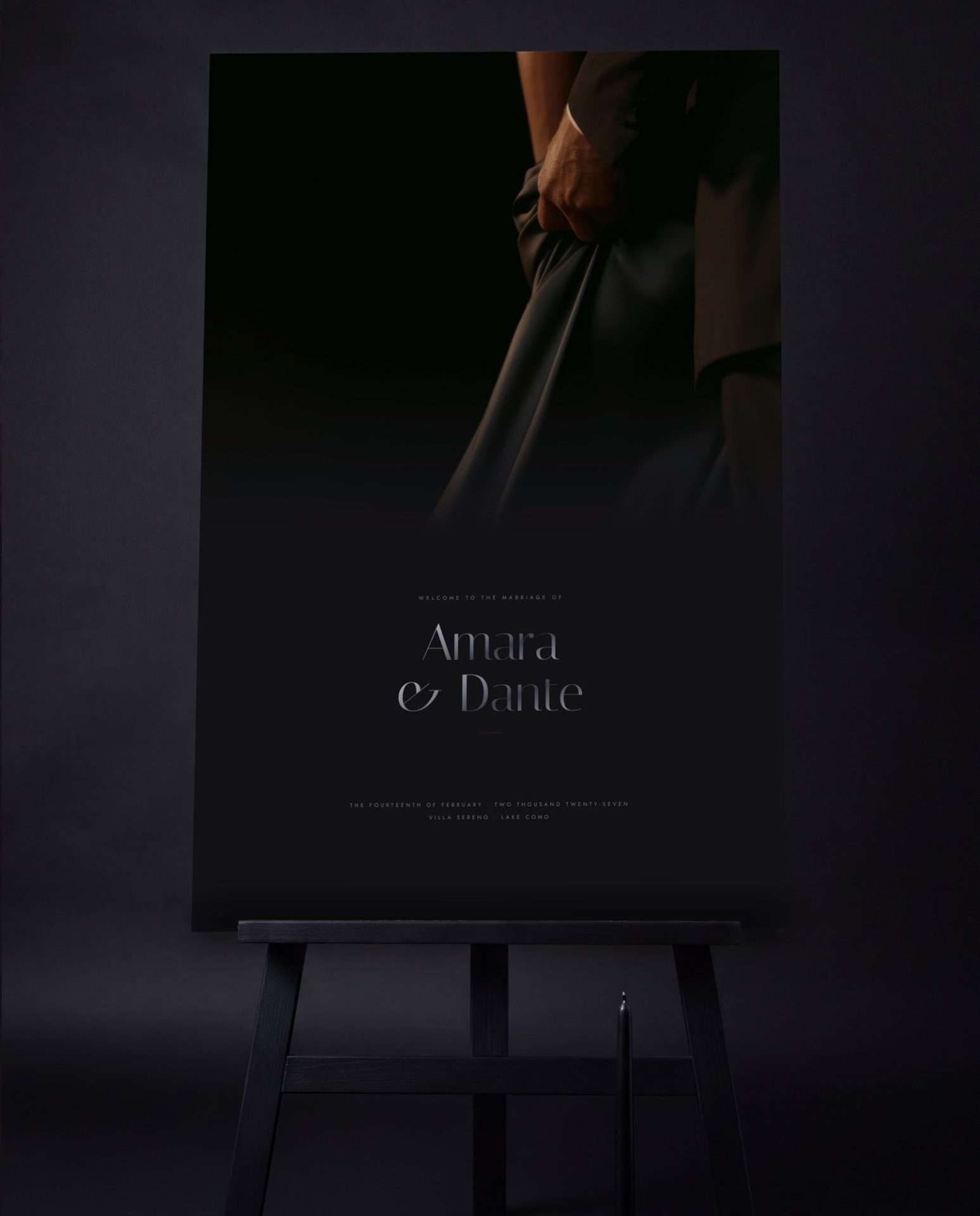 A hand resting at the waist of a black silk gown beside a dark dinner jacket, printed large format on a smoke black welcome sign