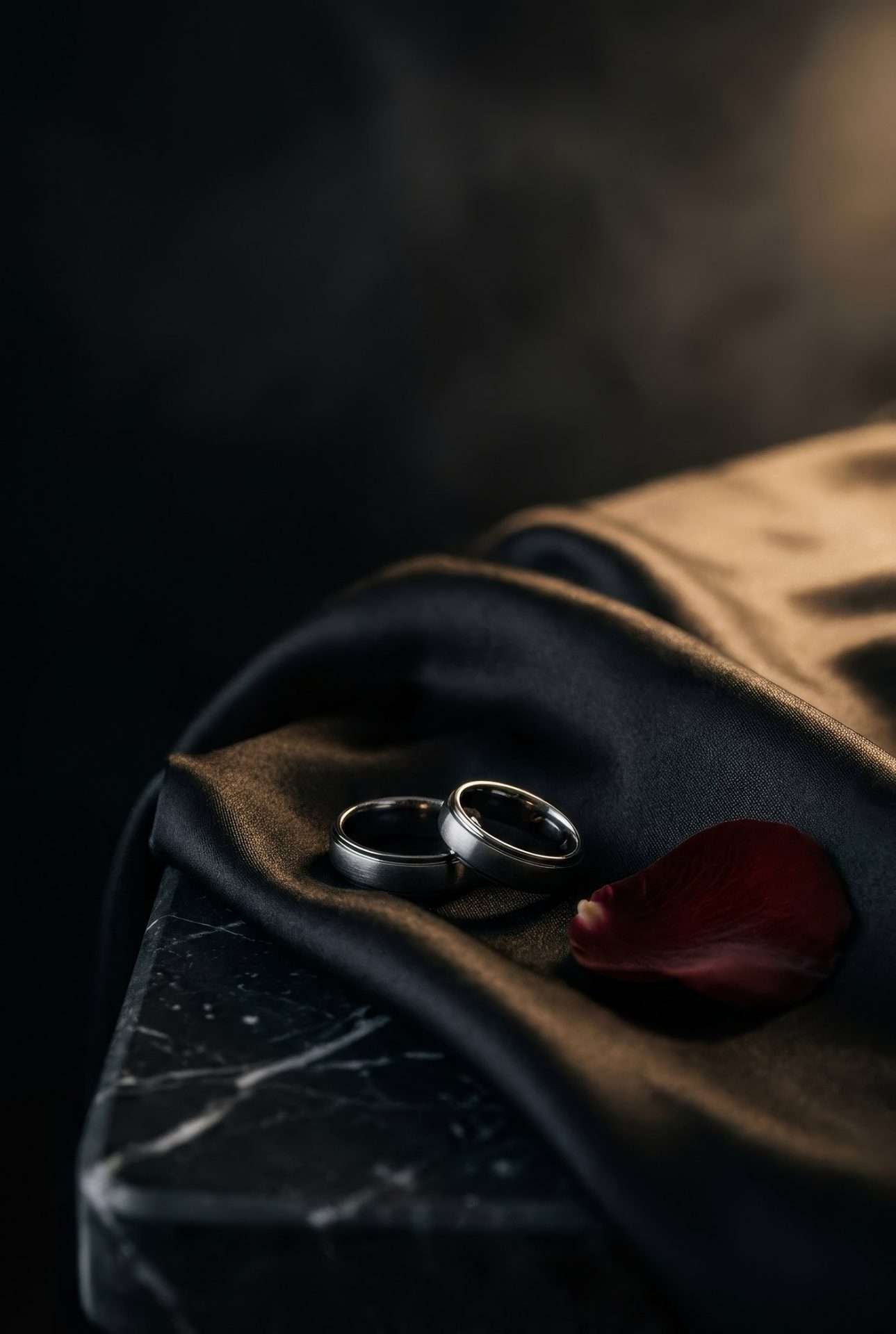 Two wedding rings on dark silk with a single petal, resting against a black marble edge